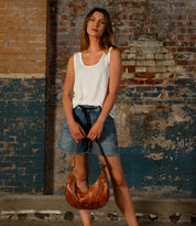A woman stands against a weathered brick wall, wearing a white tank top and denim shorts, holding the Bed|Stü Megara shoulder bag featuring chic leather details in front of her.