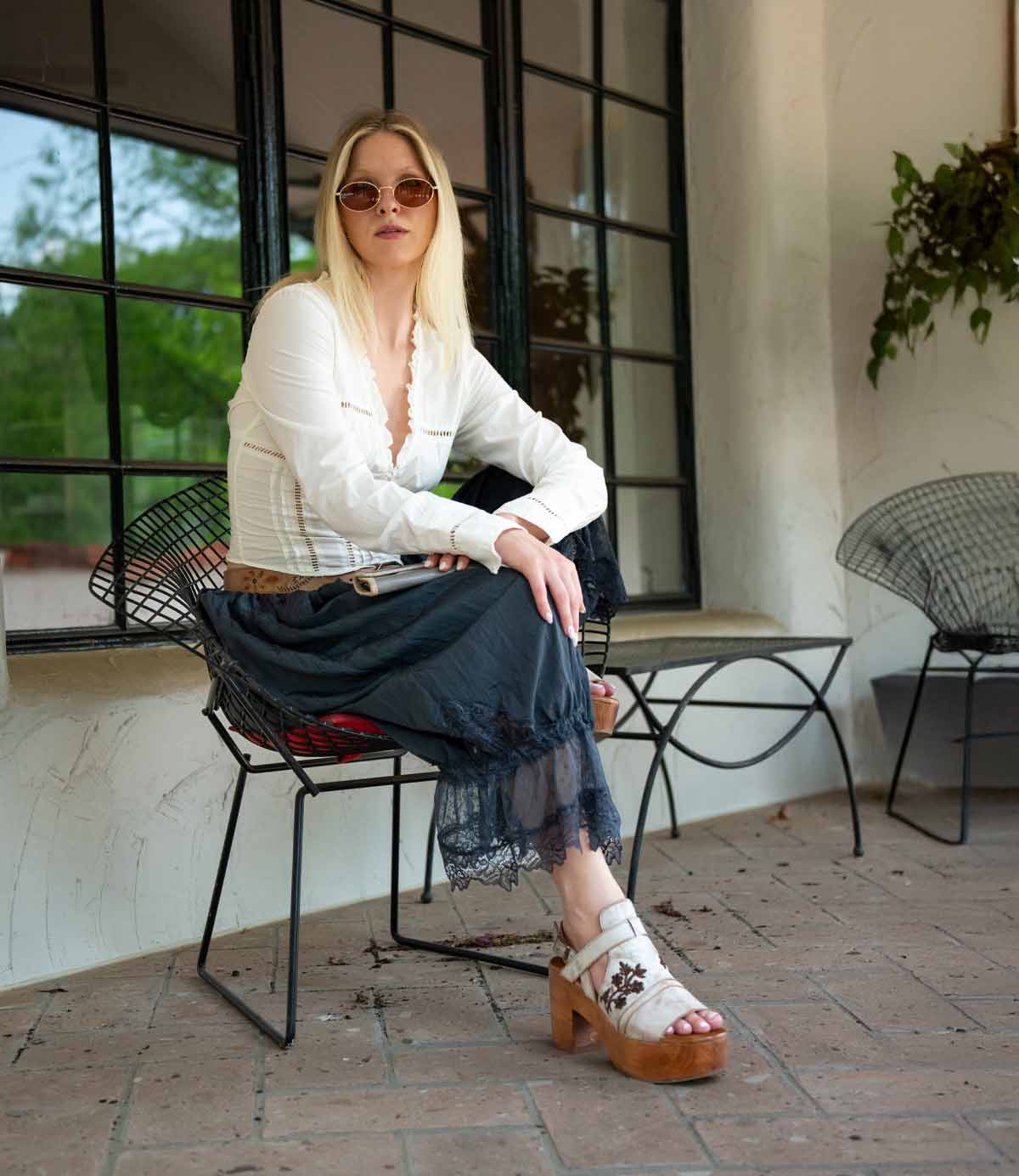 A woman with long blonde hair wears sunglasses, a white blouse, a black lace skirt, and the Bed|Stü Melodramatic platform sandals with a chunky leather heel while sitting on a black wire chair on a patio with large windows.