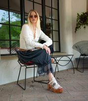 A woman with long blonde hair wears sunglasses, a white blouse, a black lace skirt, and the Bed|Stü Melodramatic platform sandals with a chunky leather heel while sitting on a black wire chair on a patio with large windows.