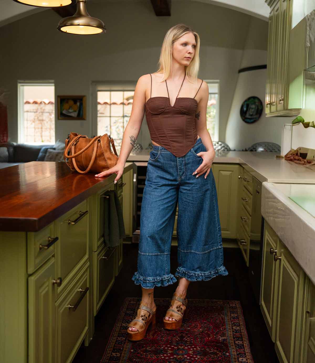 A woman stands in a kitchen wearing a brown corset top, wide-leg denim pants with ruffle hems, and the Melodramatic platform sandals by Bed|Stü, featuring a chunky leather heel. A brown handbag sits on the counter beside her.