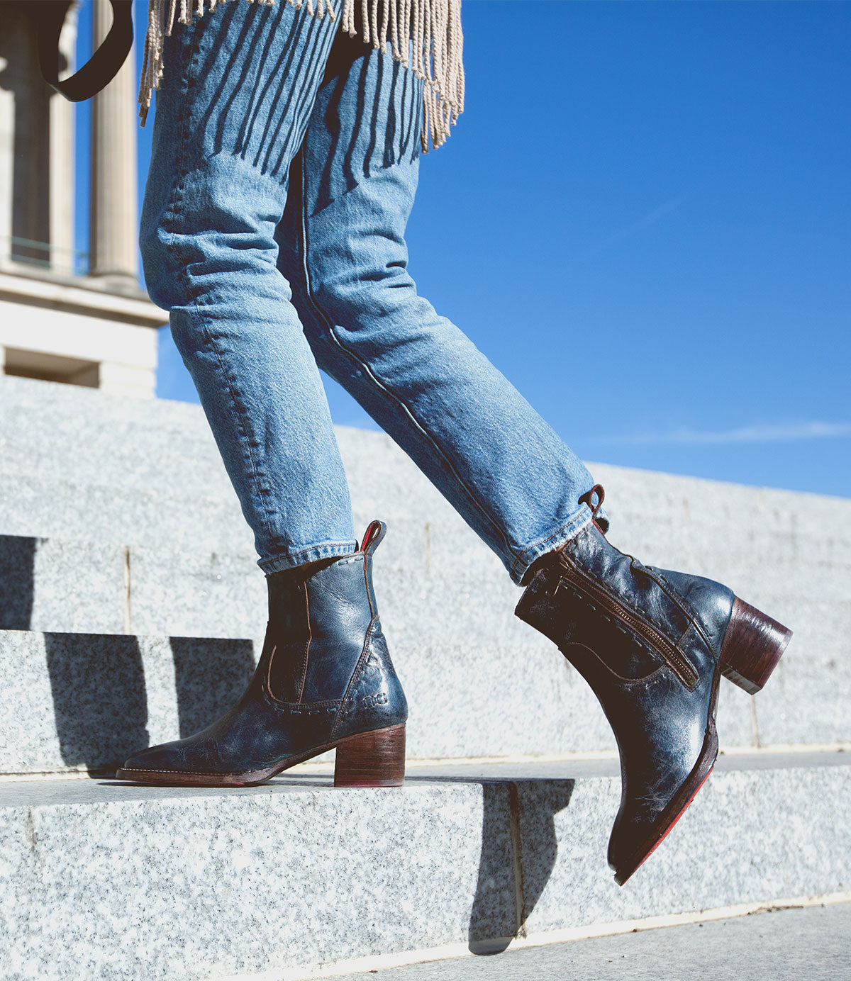 A person wearing blue jeans and the Bed|Stü Merryli boots is walking up outdoor stone steps with a clear blue sky in the background. The hand-stitched Merryli boots feature a modern square toe, adding an elegant detail to the look.