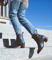 A person wearing blue jeans and the Bed|Stü Merryli boots is walking up outdoor stone steps with a clear blue sky in the background. The hand-stitched Merryli boots feature a modern square toe, adding an elegant detail to the look.
