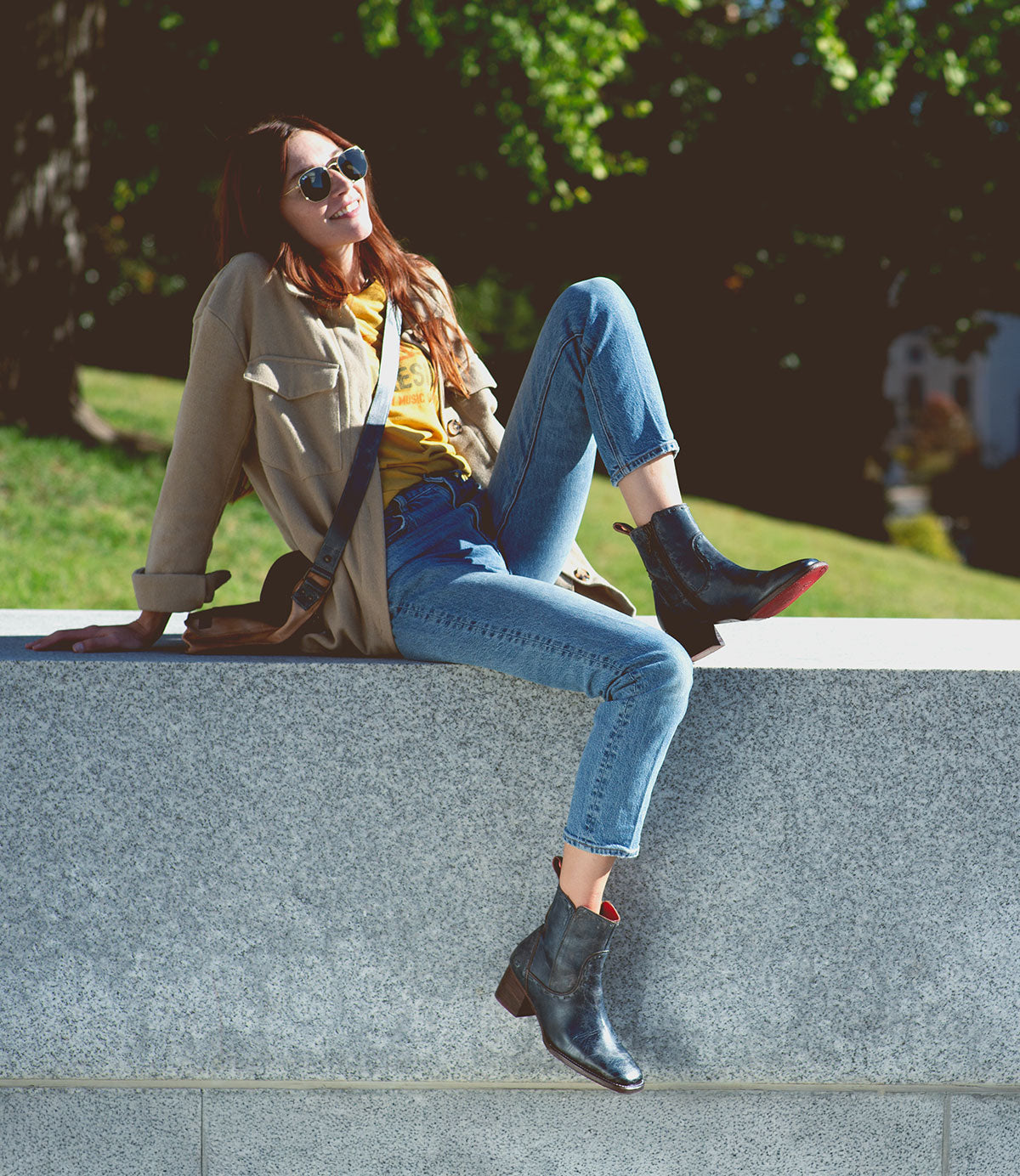 A woman wearing sunglasses and casual clothes sits on a stone ledge outdoors, smiling as she shows off her stylish Merryli leather boots by Bed|Stü, with one leg bent and the other hanging down on a sunny day.