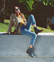A woman wearing sunglasses and casual clothes sits on a stone ledge outdoors, smiling as she shows off her stylish Merryli leather boots by Bed|Stü, with one leg bent and the other hanging down on a sunny day.