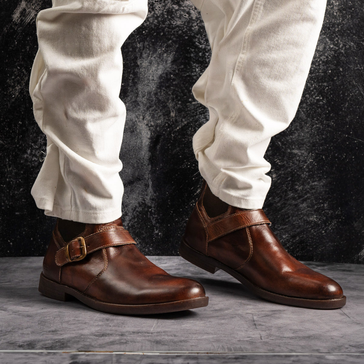 A person wearing the Bed|Stü Michelangelo brown leather dress shoes with a buckle accent and off-white pants stands on a gray textured floor with a dark background.