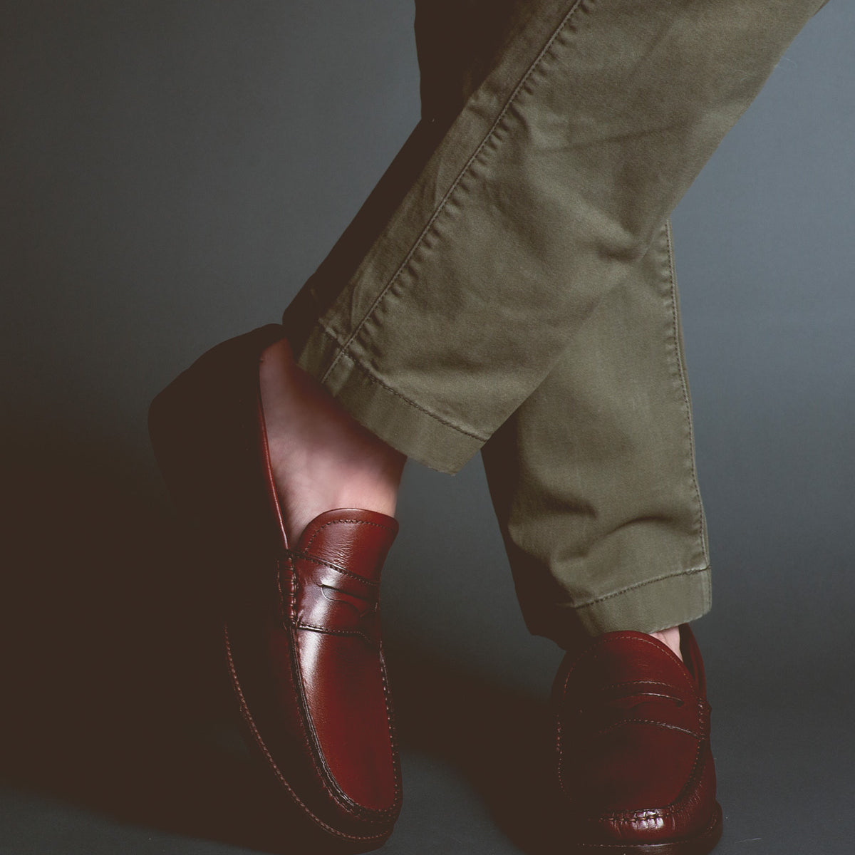 Clad in Bed|Stü’s Moat olive green pants and dark brown handcrafted loafers, a person stands with their legs crossed against a dark background.