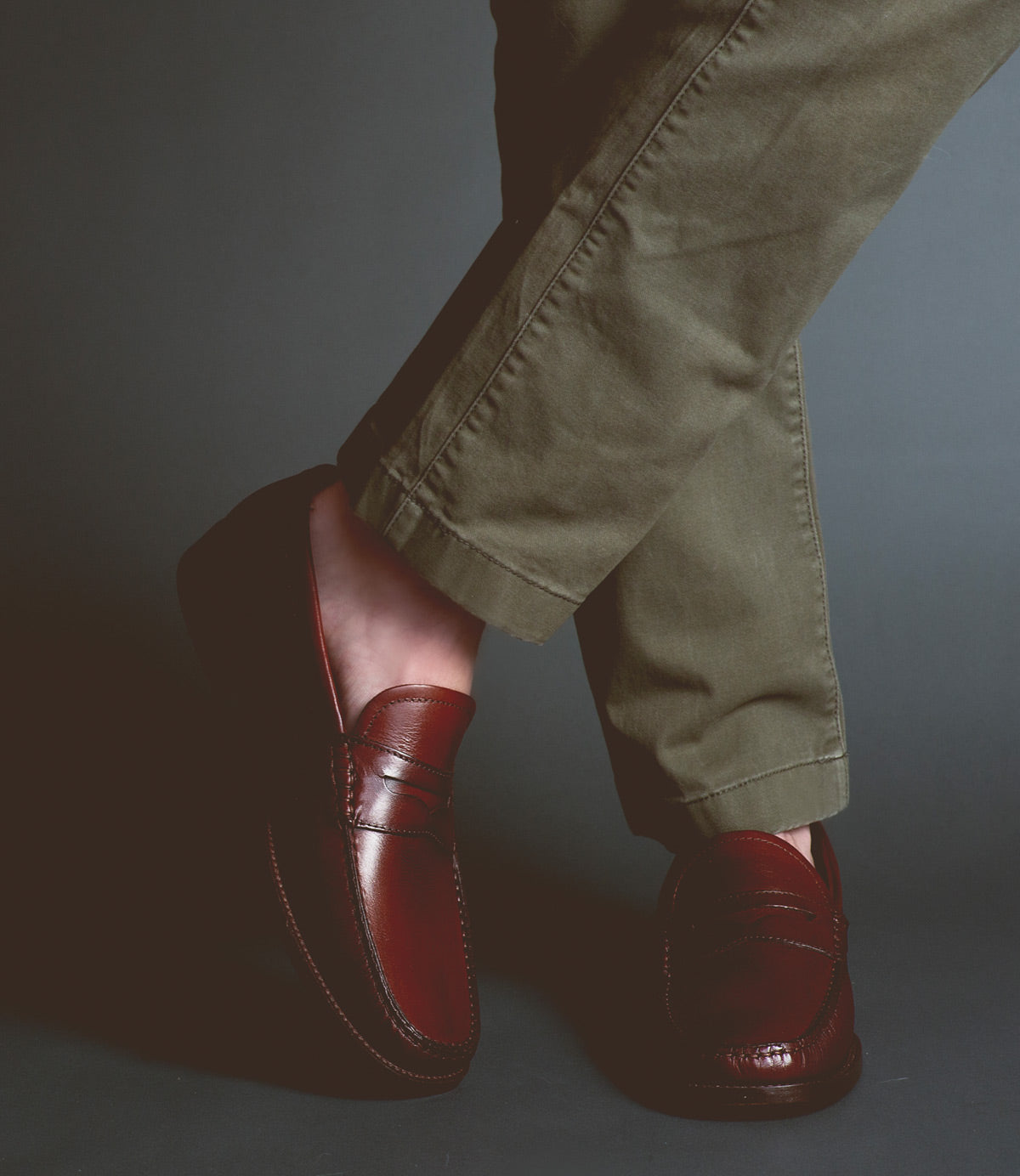 Clad in Bed|Stü’s Moat olive green pants and dark brown handcrafted loafers, a person stands with their legs crossed against a dark background.
