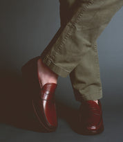 Clad in Bed|Stü’s Moat olive green pants and dark brown handcrafted loafers, a person stands with their legs crossed against a dark background.