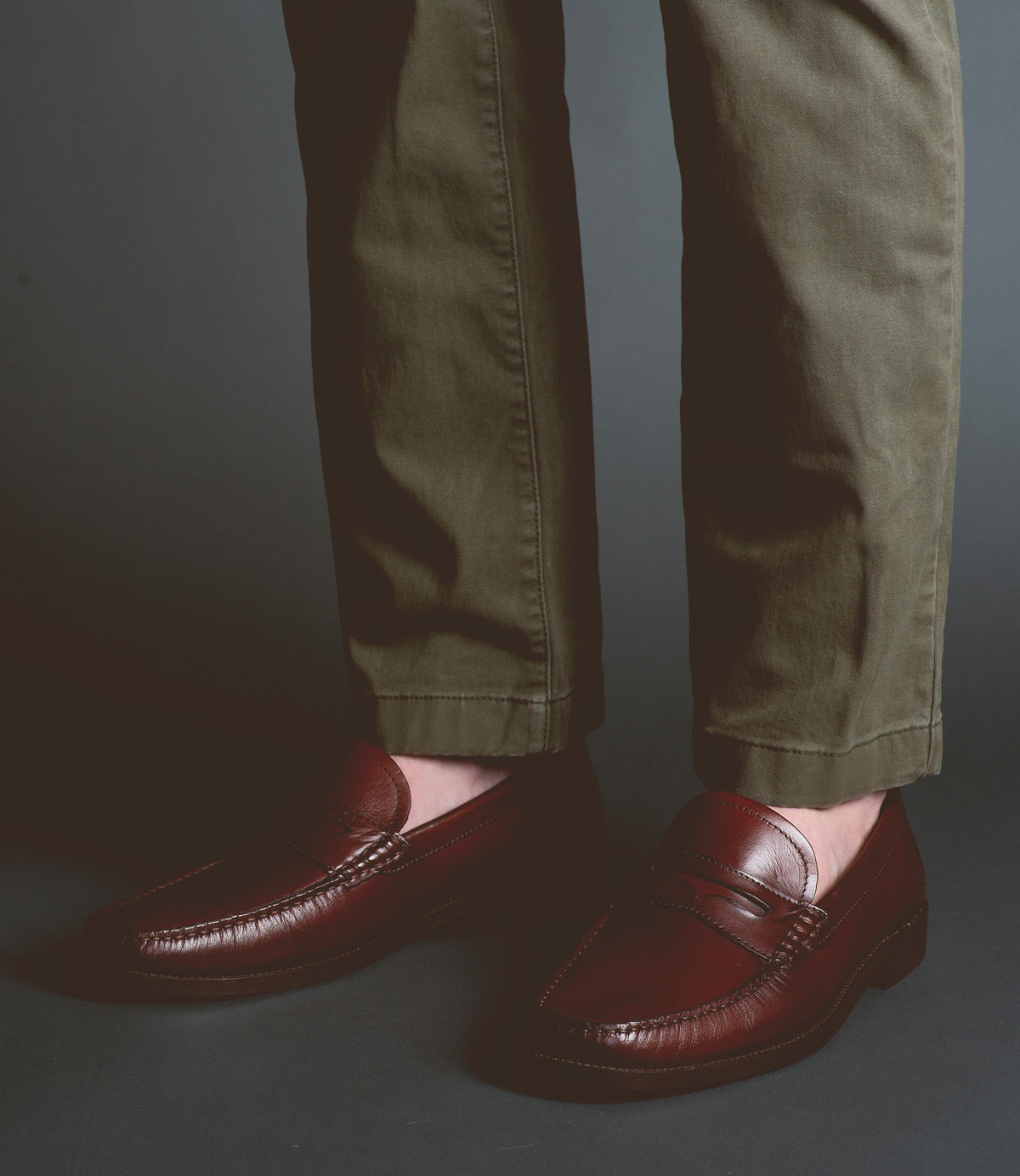 A person wearing olive green pants and a pair of Bed|Stü's Moat classic Italian leather loafers stands on a dark surface against a gray background.