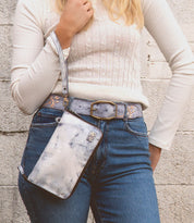 A person wearing a white sweater, blue jeans, and an embroidered floral belt holds the Mohawk pouch by Bed Stu, featuring a white and blue marble design with a wrist strap.