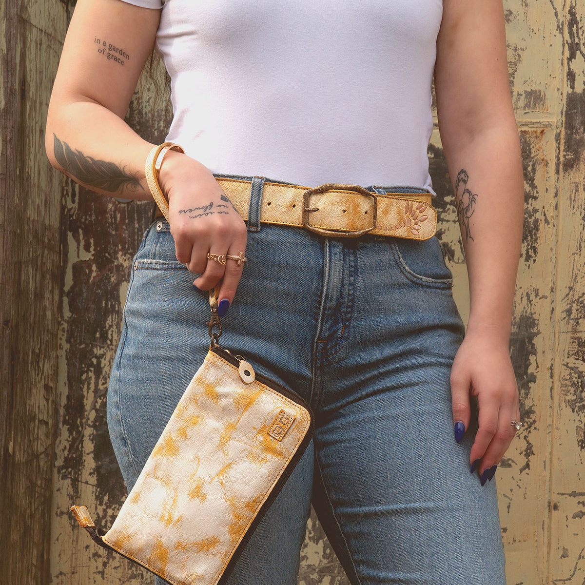 A person in a white t-shirt and blue jeans holds a yellow patterned wallet. They have tattoos on their arms and wear the Mohawk women's distressed leather belt with embroidered details by Bed Stu, perfectly matching their style.