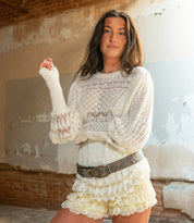 A woman stands indoors against a worn wall, wearing the Bed Stu Mohawk white knit sweater with puffy sleeves and cream lace shorts, styled with a chic women's floral belt.