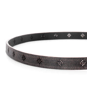 The Bed|Stü Monae II is a black leather women’s belt featuring a cracked texture and small, cutout flower-shaped patterns along its length, displayed in a curved position on a white background.