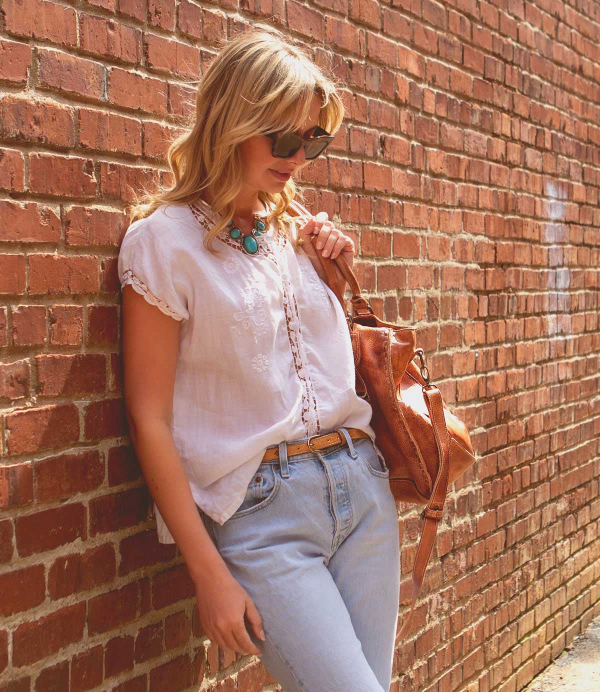 A woman in sunglasses and a white blouse leans against a brick wall, carrying the Monae II brown bag by Bed|Stü and looking down. She wears light blue jeans paired with a versatile women’s belt and a turquoise necklace.