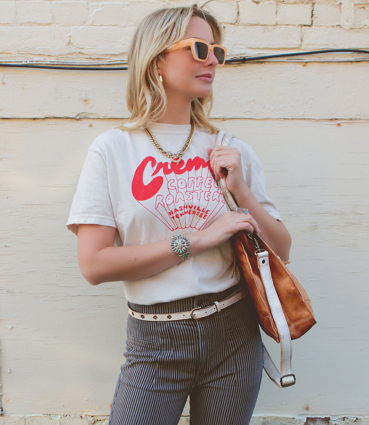 A woman wearing sunglasses, a graphic t-shirt, striped pants cinched with a versatile women’s belt, and holding the Bed|Stü Monae II brown bag stands against a light-colored wall with exposed wires near the top.