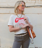A woman wearing sunglasses, a graphic t-shirt, striped pants cinched with a versatile women’s belt, and holding the Bed|Stü Monae II brown bag stands against a light-colored wall with exposed wires near the top.