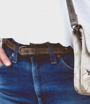 A person wearing blue jeans with a floral cutout belt, a white shirt, and carrying the Bed|Stü Monae II light-colored leather shoulder bag.
