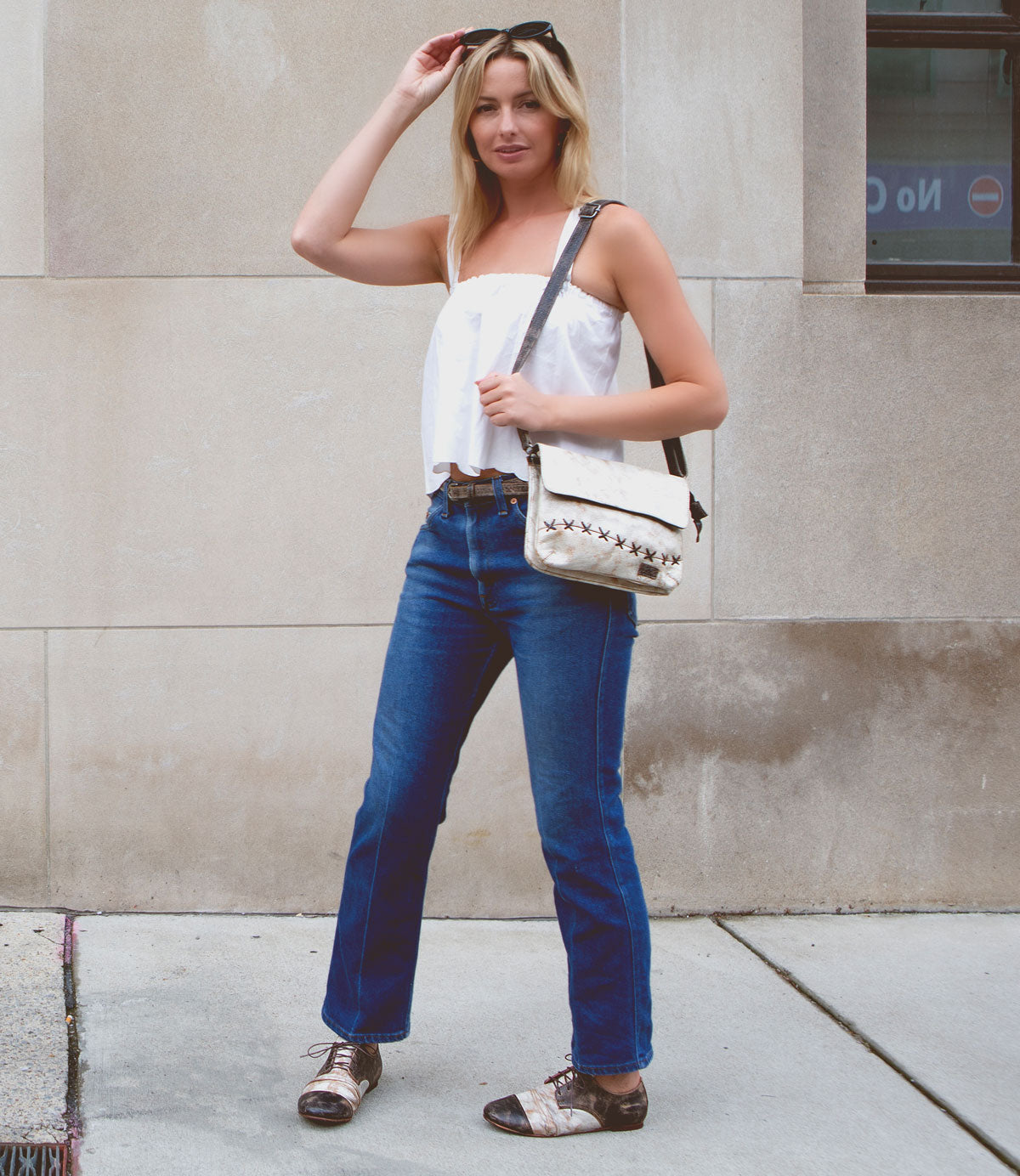 A woman stands on the sidewalk against a beige stone wall, wearing a white top, blue jeans, and the versatile Bed|Stü Monae II women’s leather belt; she holds sunglasses and a white shoulder bag, and completes her look with patterned shoes.