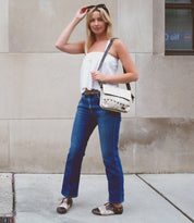 A woman stands on the sidewalk against a beige stone wall, wearing a white top, blue jeans, and the versatile Bed|Stü Monae II women’s leather belt; she holds sunglasses and a white shoulder bag, and completes her look with patterned shoes.
