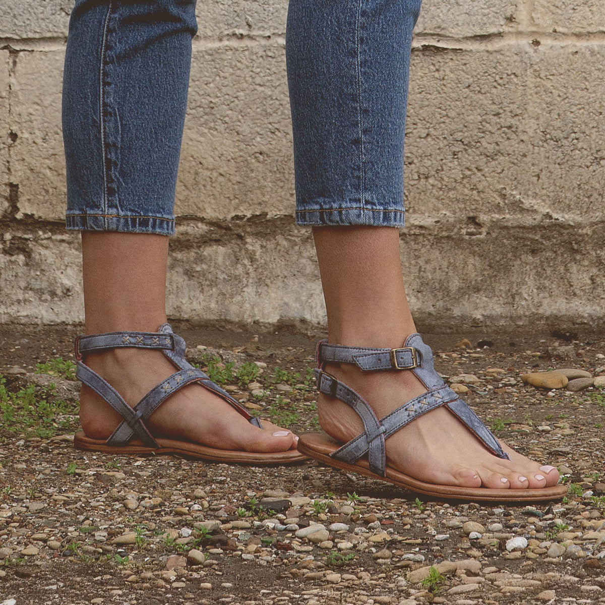 A person wearing the Bed|Stü "Moon" blue strappy leather sandals with a cushioned insole and cropped jeans stands on gravel ground beside a weathered concrete wall.