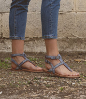 A person wearing the Bed|Stü "Moon" blue strappy leather sandals with a cushioned insole and cropped jeans stands on gravel ground beside a weathered concrete wall.