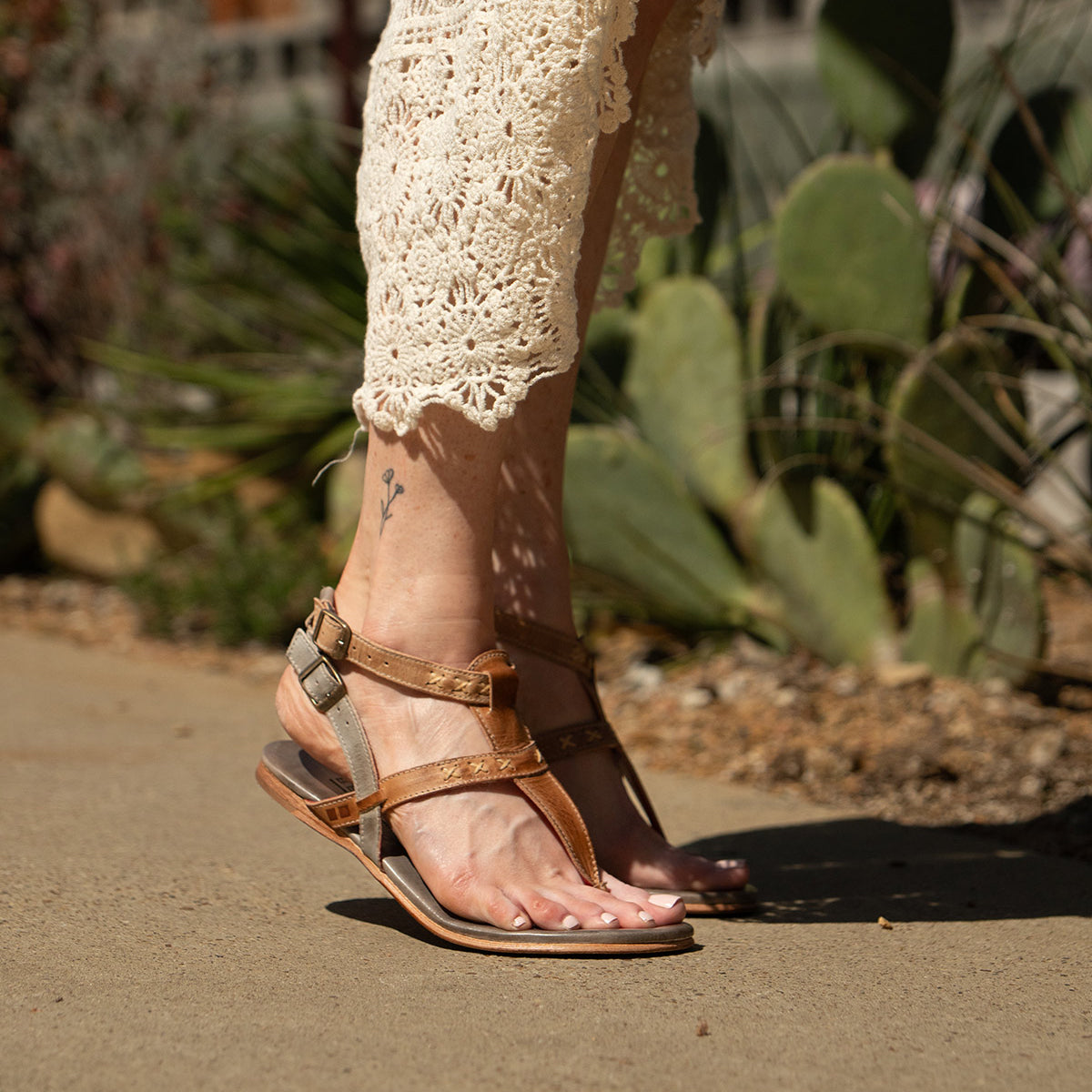 A person wearing the "Moon" brown leather sandals with an ankle buckle by Bed|Stü and a cream crochet dress walks on a sidewalk with green plants in the background.