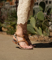 A person wearing the "Moon" brown leather sandals with an ankle buckle by Bed|Stü and a cream crochet dress walks on a sidewalk with green plants in the background.
