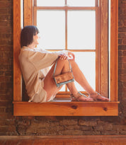 A person in a light outfit sits on a wooden windowsill, looking out the window with a small tan purse resting on their knees, their feet comfortably slipped into Bed|Stü’s Moon leather sandals, featuring an elegant ankle buckle.