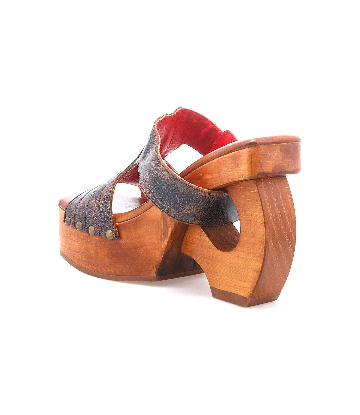 The Bed|Stü Mutable is a wooden platform sandal with a chunky, curved heel and vintage influence, featuring a dark leather upper, decorative buckle detail, brass rivets, and red inner lining shown on a white background.