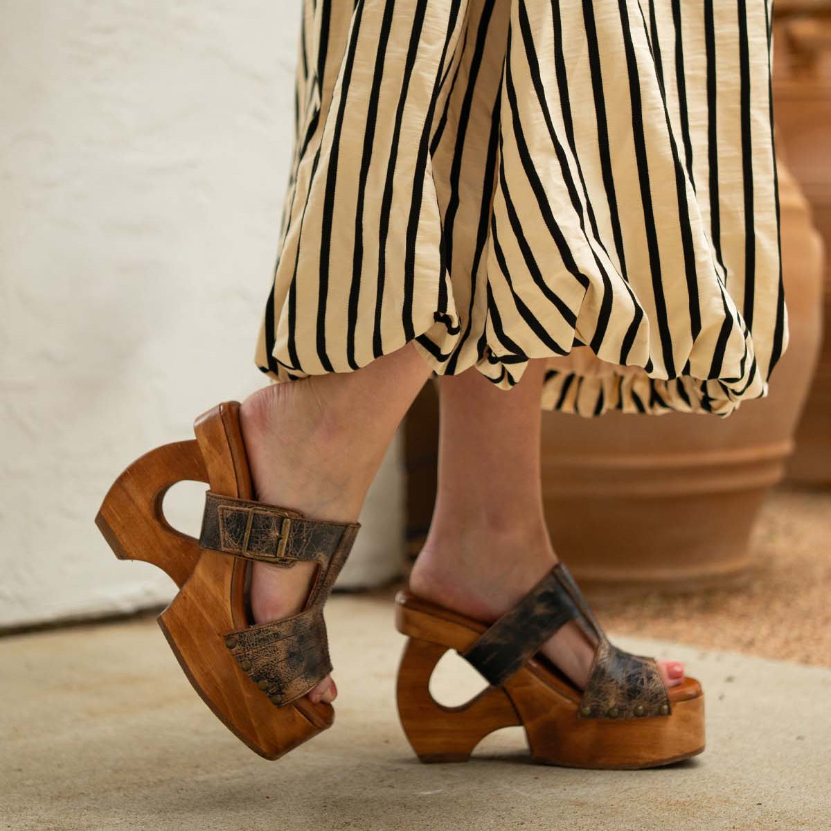 Wearing the Bed|Stü Mutable chunky wooden platform sandals, featuring cut-out heels and a decorative buckle, this look pairs perfectly with a beige and black striped skirt for a subtly vintage-inspired style.