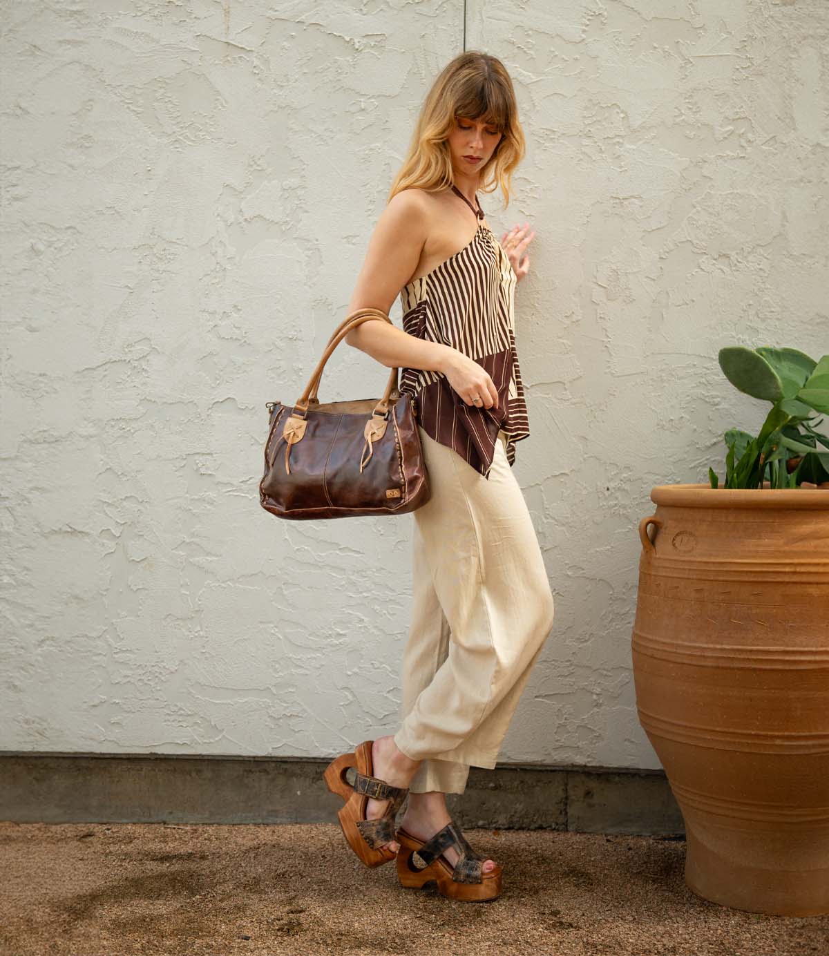 A woman stands near a large planter against a pale textured wall, carrying the Bed|Stü Mutable brown handbag while wearing a striped tank top, cream pants, and wooden platform heel sandals with a decorative buckle.