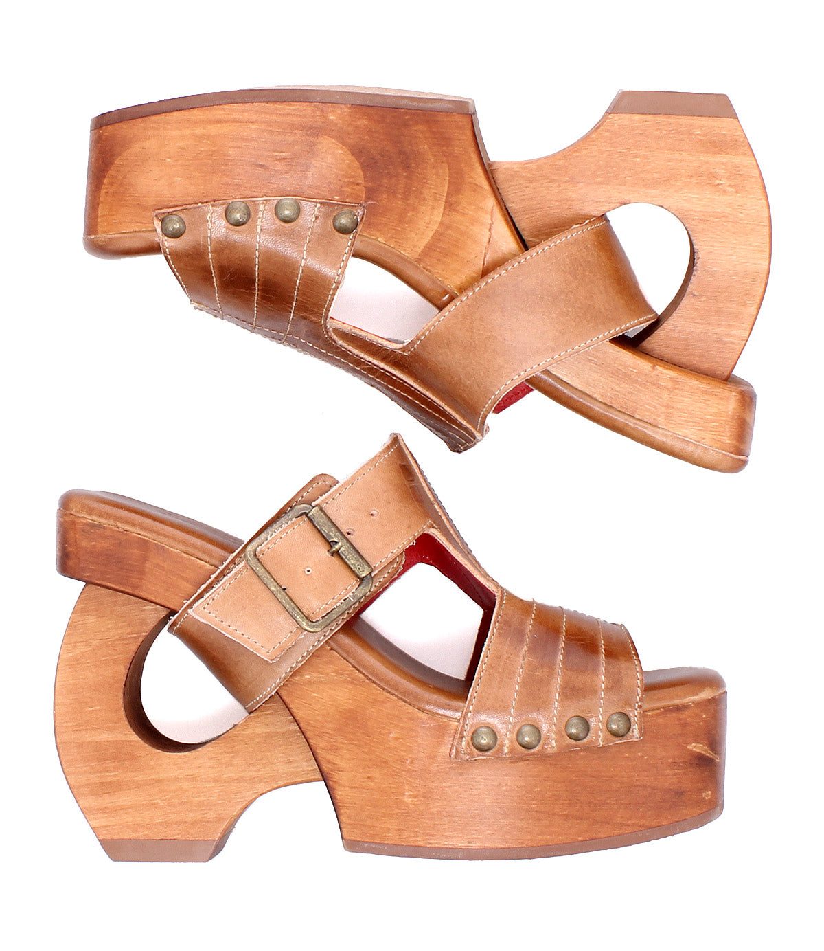 A pair of Mutable sandals by Bed|Stü, crafted from brown leather and wood with platform heels, featuring a cutout heel, decorative buckle strap, and metal rivet accents, are displayed side by side on a white background and radiate a subtle vintage vibe.
