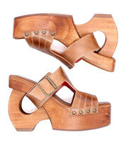 A pair of Mutable sandals by Bed|Stü, crafted from brown leather and wood with platform heels, featuring a cutout heel, decorative buckle strap, and metal rivet accents, are displayed side by side on a white background and radiate a subtle vintage vibe.
