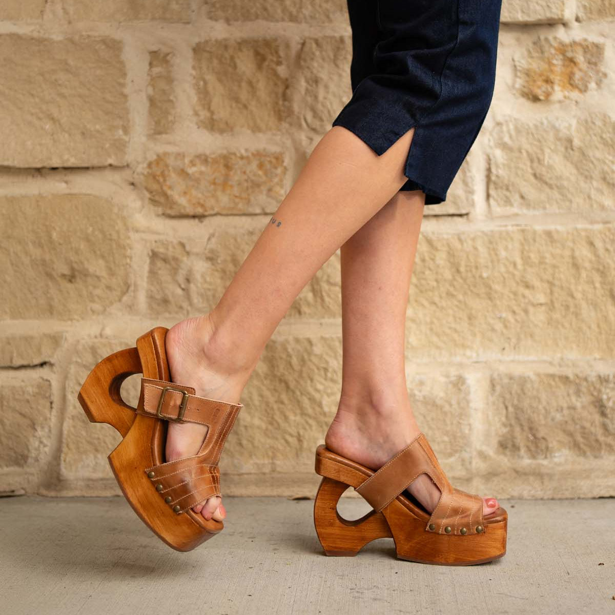 Wearing Bed|Stü's Mutable sandals—tan wooden platform heels adorned with a decorative buckle—a person in dark cropped pants stands on a concrete surface before a beige stone wall, channeling a subtle vintage vibe.