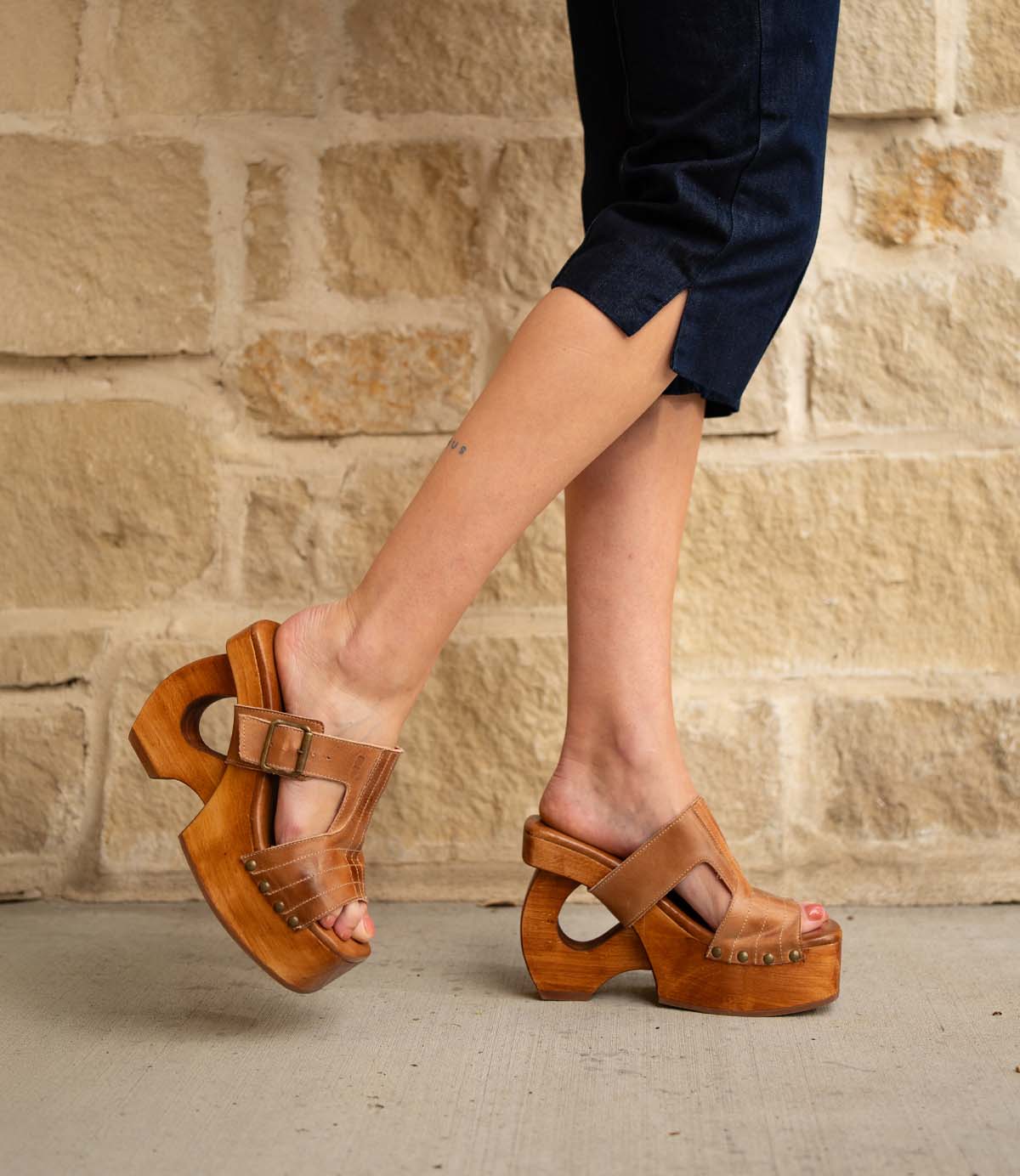 Wearing Bed|Stü's Mutable sandals—tan wooden platform heels adorned with a decorative buckle—a person in dark cropped pants stands on a concrete surface before a beige stone wall, channeling a subtle vintage vibe.