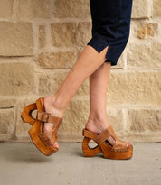 Wearing Bed|Stü's Mutable sandals—tan wooden platform heels adorned with a decorative buckle—a person in dark cropped pants stands on a concrete surface before a beige stone wall, channeling a subtle vintage vibe.