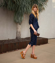 Wearing a dark blue outfit and the Bed|Stü Mutable wooden platform sandals with a decorative buckle, a woman with long hair walks on a sidewalk, carrying a small brown purse. A palm tree and light-colored wall in the background enhance the look’s subtle vintage vibe.