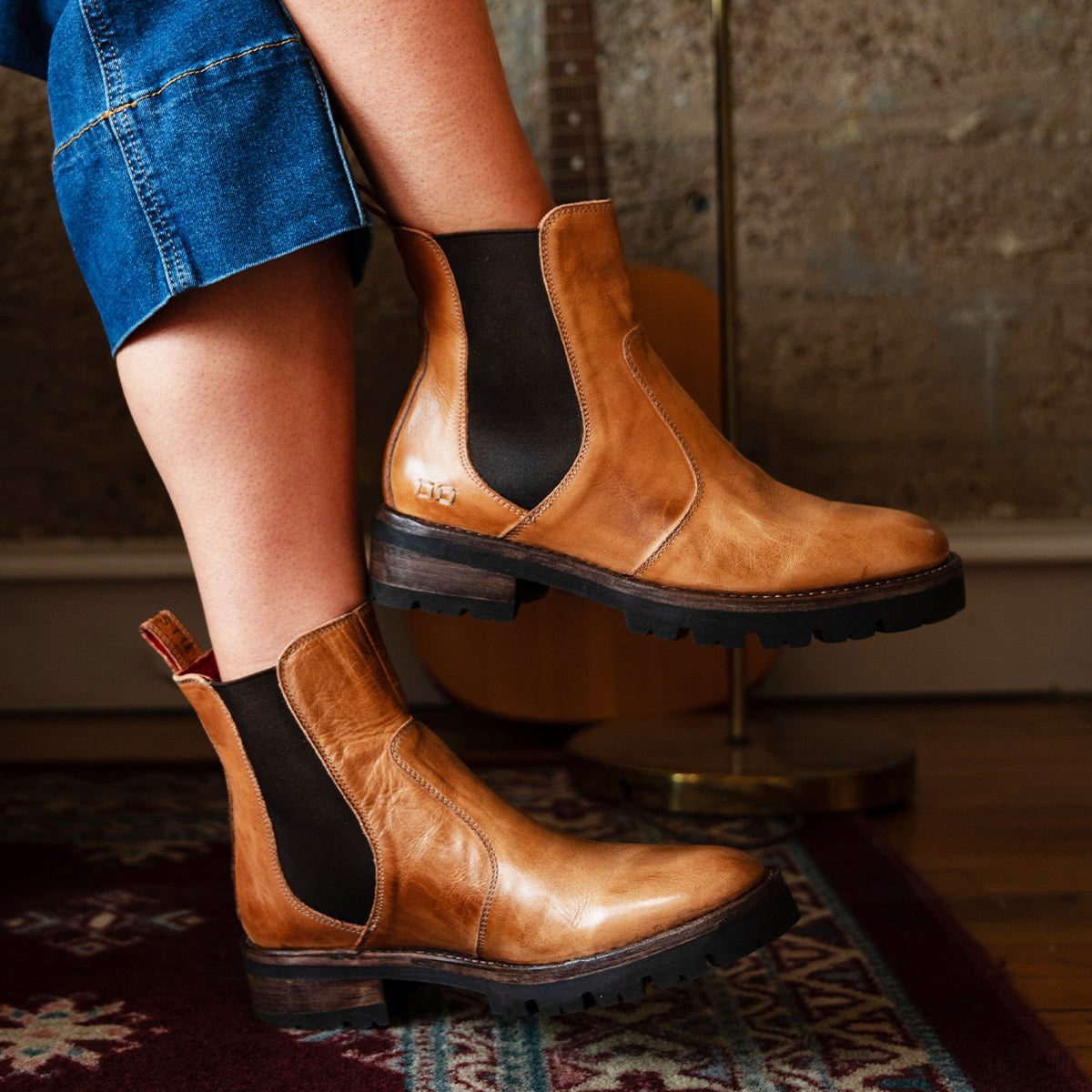A person wearing Bed|Stü Nandi III brown leather Chelsea boots with dual side gores and blue jeans sits on a patterned rug, with a guitar and textured wall in the background, showcasing timeless style.