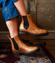 A person wearing Bed|Stü Nandi III brown leather Chelsea boots with dual side gores and blue jeans sits on a patterned rug, with a guitar and textured wall in the background, showcasing timeless style.