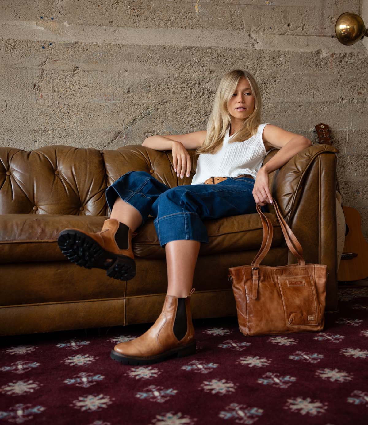 A woman sits on a brown leather sofa, exuding a refined look in her white top, blue jeans, and timeless Bed|Stü Nandi III brown Chelsea boots. A matching brown bag rests on the sofa, while a guitar leans against the wall in the background.