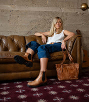 A woman sits on a brown leather sofa, exuding a refined look in her white top, blue jeans, and timeless Bed|Stü Nandi III brown Chelsea boots. A matching brown bag rests on the sofa, while a guitar leans against the wall in the background.