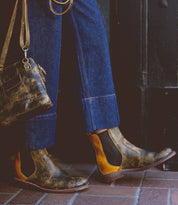 A person wearing Bed|Stü's Nandi distressed brown leather Chelsea boots with a classic slip-on design, paired with blue cuffed jeans and carrying a matching brown leather bag, stands on a brick sidewalk.