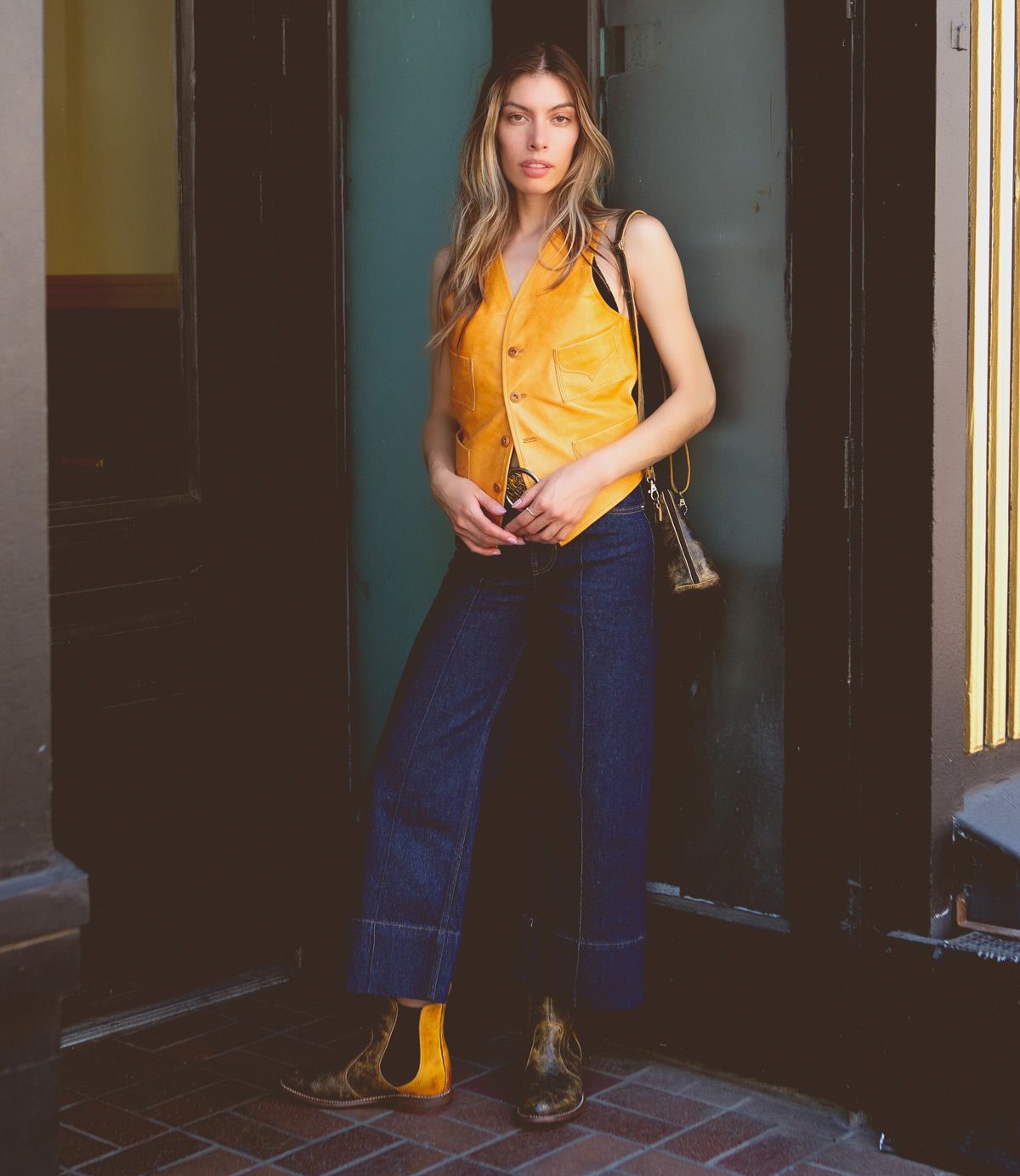 A woman stands in a doorway wearing a yellow vest, dark wide-leg jeans, and versatile Bed|Stü Nandi Chelsea boots with a slip-on design, while holding a small shoulder bag.