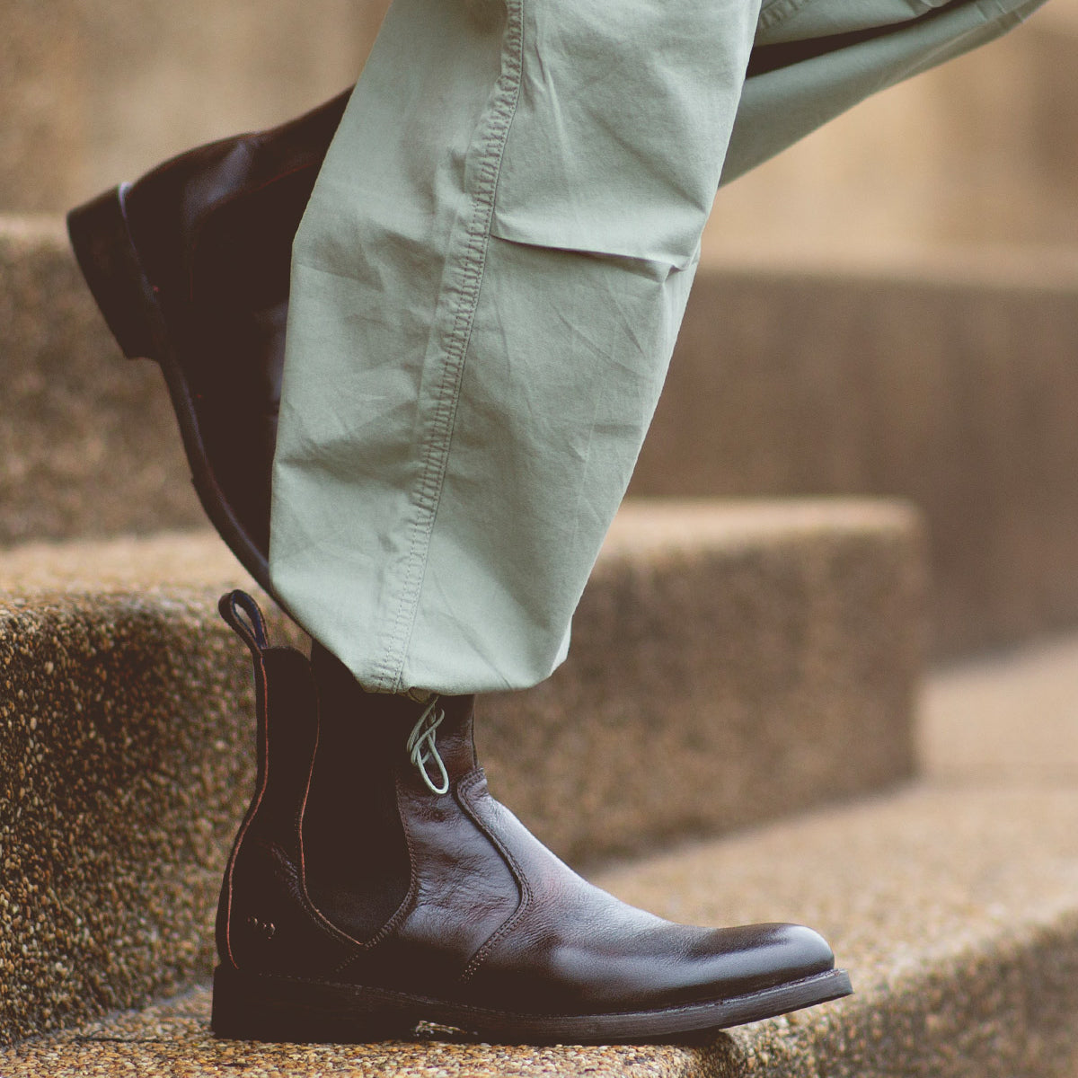 A person wearing light green pants and a pair of Bed|Stü Nando dark brown hand-worked leather Chelsea boots stands on stone steps.