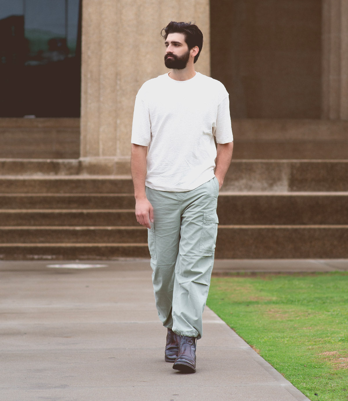 A bearded man wearing a white shirt, light green cargo pants, and gray Bed|Stü Nando Goodyear welt Chelsea boots walks on a pavement near stone steps and grass.
