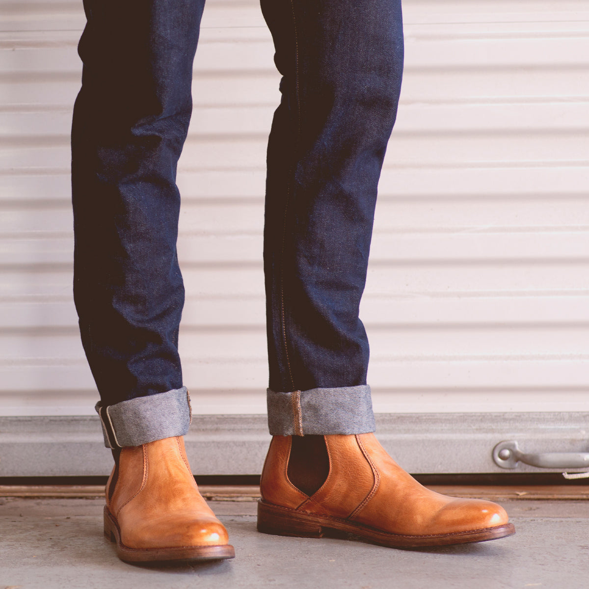 A person wears cuffed dark blue jeans and tan Bed|Stü Nando Chelsea boots, crafted from hand-worked leather, while standing on a concrete floor in front of a white garage door.