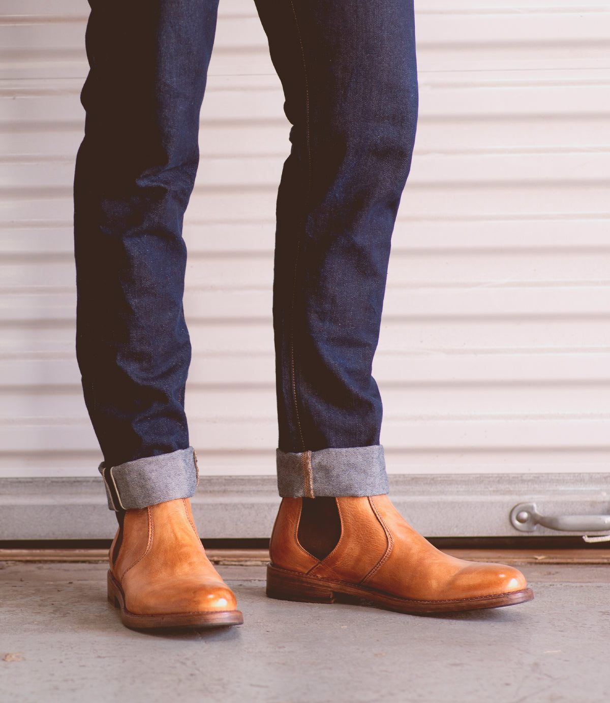 A person wears cuffed dark blue jeans and tan Bed|Stü Nando Chelsea boots, crafted from hand-worked leather, while standing on a concrete floor in front of a white garage door.