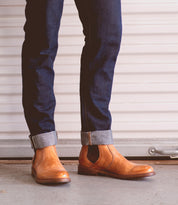 A person wears cuffed dark blue jeans and tan Bed|Stü Nando Chelsea boots, crafted from hand-worked leather, while standing on a concrete floor in front of a white garage door.