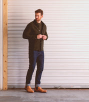 A man stands indoors in front of a closed white garage door, wearing a dark jacket, blue jeans with rolled cuffs, and brown Bed|Stü Nando Chelsea boots crafted from hand-worked leather.