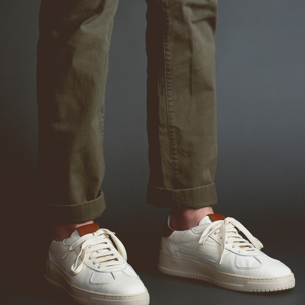 Wearing olive green pants and classic men’s footwear—the sleek "North" white sneakers by Bed|Stü—a person stands against a dark background.